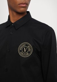 Camicia nera in cotone con bottoni, caratterizzata da un logo Versace Jeans Couture ricamato in oro sul lato sinistro del petto. Collo classico e texture liscia.