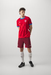 Rød fodboldtrøje med blå detaljer og emblem, sammen med maroon shorts, hvide sokker og sorte støvler. Moderne pasform og tekstureret stof.