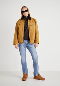 Chaqueta de sarga marrón con dos bolsillos en el pecho sobre un suéter oscuro, combinada con jeans de mezclilla azul claro y zapatillas marrones. El modelo lleva gafas de sol negras.