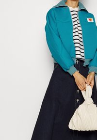 Turkos zip-up bomberjacka med en ljusblå krage, randig skjorta under och en krämfärgad plisserad handväska. Mörk denimkjol med knappar.