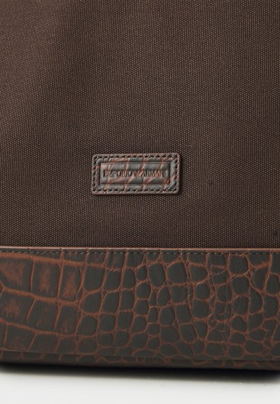 Brown fabric bag featuring a textured dark brown leather base, with a metallic Emporio Armani logo plaque attached.