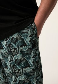 Broek met een zwarte basiskleur en een groen en teal bladerenpatroon. Gemaakt van zachte stof met een elastische tailleband en zijzak.