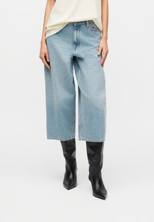 Femme portant un jean large et court bleu clair, un haut crème et des bottes noires à bout pointu montant jusqu'aux genoux sur fond blanc.