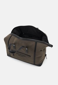 Sac de voyage en toile vert olive avec des accents en cuir noir, quincaillerie en argent, poignées doubles et fermeture éclair. L'intérieur comprend une poche zippée.