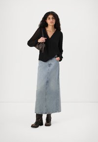 Svart bluse med gjennomsiktige ermer, V-hals, sammen med et langt, lyseblått denimskjørt og mørkebrune ankelstøvler. Holder en liten brun veske.