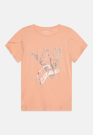 Koszula z krótkim rękawem w kolorze brzoskwiniowym z błyszczącym srebrnym trójkątnym logo wypełnionym kryształkami oraz napisem „Guess” w srebrnym stylu pisma.