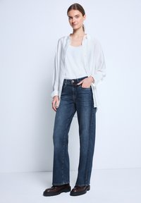 Jeans de mezclilla azul oscuro con un corte relajado, combinados con una camiseta sin mangas blanca y una camisa blanca transparente abierta, complementados con botas gruesas marrones.