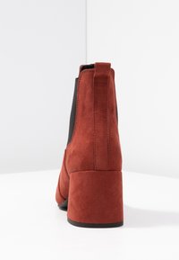 Botte de cheville en daim rouge avec un talon bloc et un panneau latéral élastique noir, photographiée de l'arrière sur une surface blanche.