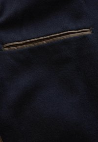 Marinblå ylletyg med en slät textur som har en horisontell dragkedjeficka kantad i mörkbrunt. Enkel design med minimalistiska accenter.
