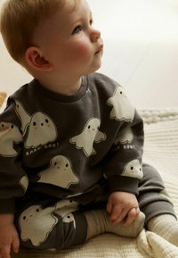 Body gris avec des motifs de fantômes ludiques, doté d'un col rond et de poignets côtelés. Enfant assis les mains jointes sur une couverture texturée.