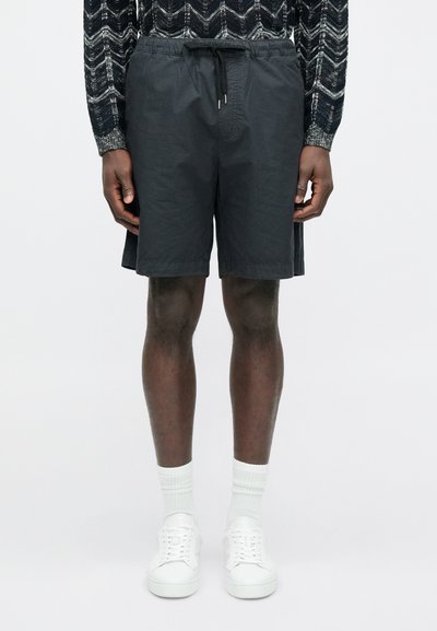 Czarne szorty z tkaniny z gumką w talii i bocznymi kieszeniami, cechujące się lekką fakturą i luźnym krojem. Połączone z białymi trampkami i skarpetkami.