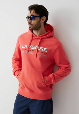 Mężczyzna w koralowej bluzie z kapturem z napisem „DIVERSE NAUTICAL CLUB” i niebieskich okularach przeciwsłonecznych, ręce w kieszeniach, oparty o białą ścianę.