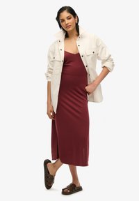 Vestido midi ajustado en color burdeos, textura suave, combinado con una chaqueta de botones de gran tamaño en color blanco roto y sandalias marrones de dedo. Sin patrones visibles.
