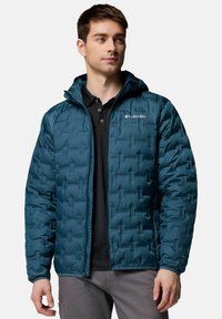 Turkusowa kurtka ocieplana z kapturem, z pikowanym wzorem, na zamek błyskawiczny oraz logo Columbii po lewej stronie klatki piersiowej. Nosi się ją na czarnym polo.