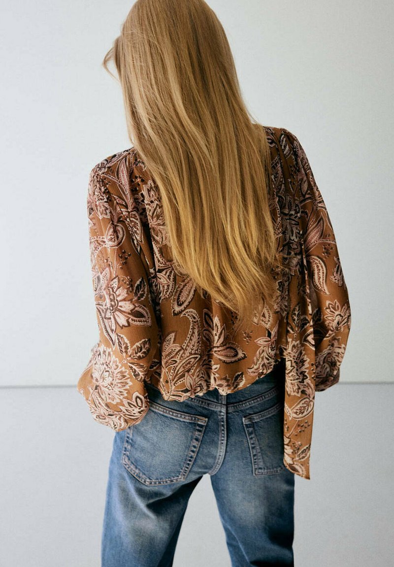 Personne aux longs cheveux blonds portant un chemisier marron à motifs et un jean bleu, debout de dos sur un fond uni.