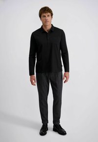 Schwarzes Langarm-Polo-Shirt mit Kragen und Brusttasche, kombiniert mit strukturierten grauen Hosen und schwarzen Schuhen, stehend vor einem einfarbigen Hintergrund.