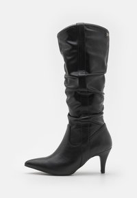 Black knee-high boot made of smooth leather, featuring a pointed toe, slouched design, and a moderate heel.  Accent on the upper side.