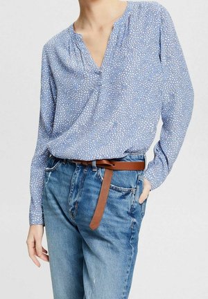 Blaue gemusterte Bluse mit V-Ausschnitt, langen Ärmeln und gerafften Details. Kombiniert mit hochgeschnittenen, verwaschenen blauen Jeans und einem braunen Ledergürtel.