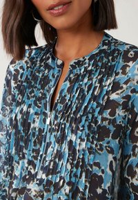 Sininen ja musta paita, jossa on leopardikuosiset, pleatatut yksityiskohdat ja vetskari kaula-aukossa. Tasainen kangas ja rento istuvuus.