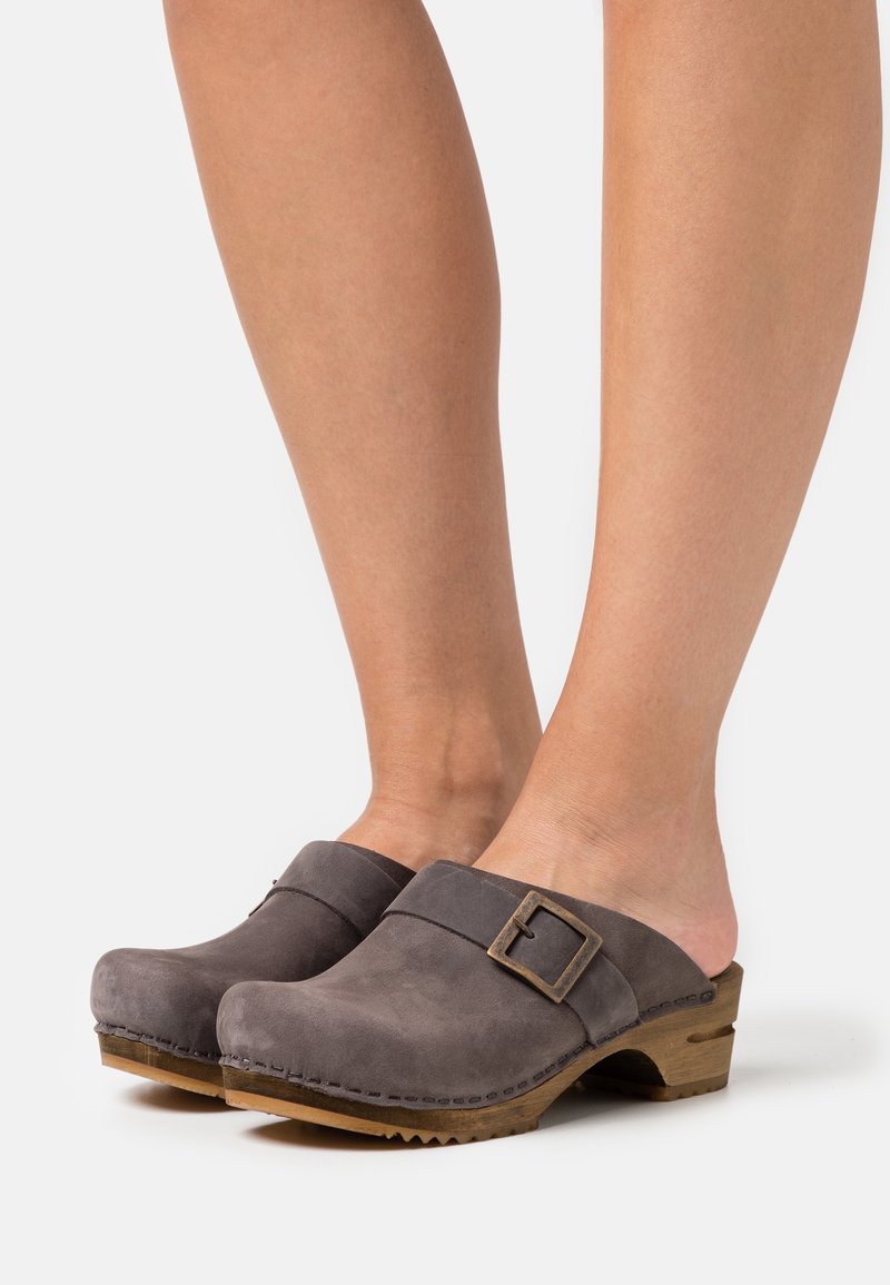 Pieds portant des sabots en daim gris avec semelles en bois et une boucle latérale, présentés sur un fond clair uni.