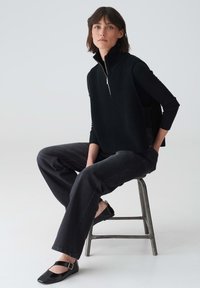 Frau, die auf einem Metallhocker sitzt und einen schwarzen ärmellosen Reißverschluss-Pullover, ein schwarzes Langarmoberteil, dunkle Jeans und schwarze Mary-Jane-Schuhe trägt.