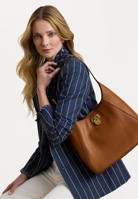 Barna bőr válltáska arany kiegészítőkkel, sima felülettel és ívelt formával. A modellen kék csíkos zakó van, ülő pózban.