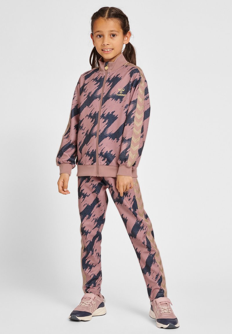 Jeune fille debout en survêtement rose et noir à motifs avec des bandes beiges en chevron et des baskets roses, souriant à la caméra.