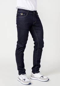 Dunkelblaue Denim-Jeans mit Slim Fit, die sichtbare Nähte und vordere Taschen aufweisen. Kombiniert mit weißen und grauen Sneakers.