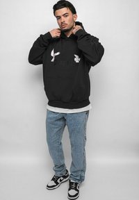 Black hoodie with white dove graphics, front kangaroo pocket, layered over a gray shirt, paired with light blue jeans and black sneakers.