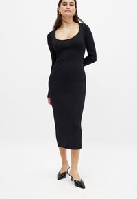 Musta ribbed pitkähihaine mekko, jossa on neliönmuotoinen pääntie, vartalonmyötäinen siluetti ja midi-pituus. Yhdistetty mustiin kärkitossuihin.