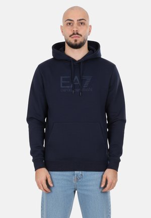 Uomo che indossa una felpa con cappuccio blu navy con il logo EA7 Emporio Armani, jeans azzurri chiari, in piedi davanti a uno sfondo grigio chiaro uniforme.
