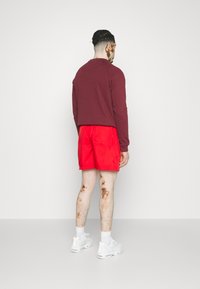 Bordová dlhá košeľa s okrúhlym výstrihom, spárovaná s jasne červenými šortkami. Biely športový obuv a viditeľné detaily textúry pokožky.