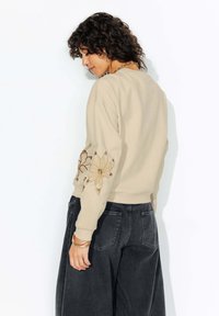 Sweatshirt beige avec design floral brodé sur la manche gauche, poignets côtelés et coupe décontractée. Associé à un pantalon large sombre.