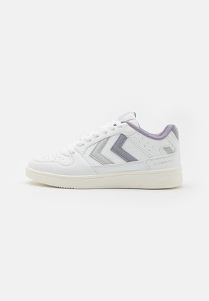 Zapatilla blanca con acentos en morado claro, detalles perforados y suela plana color crema. Presenta un diseño de chevron prominente a los lados.
