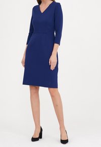 Femme debout portant une robe bleu foncé, longue jusqu’aux genoux, à manches longues, avec un col en V et des chaussures à talons noirs sur un fond blanc uni.