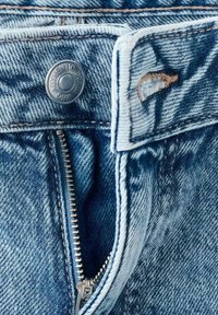 Jean en denim bleu clair avec un bouton métal marqué "AUTHENTIC DENIM" et une fermeture éclair argentée avec un rabat en tissu.