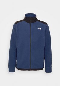 Sininen fleece-takki mustilla hartioilla, täysimittainen vetoketju edessä ja "The North Face" -logo vasemmassa rinnassa.