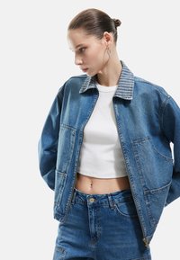 Jeansjacke mit gestreiftem Kragen, zwei Taschen vorne, mit Reißverschluss und einem leicht übergroßen Schnitt. Getragen über einem weißen, cropped Shirt.