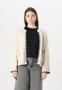 Beige Strickjacke mit schwarzem gewelltem Saum entlang der Kanten und Taschen, mit lockerer Passform und gerippten Bündchen. Getragen über einem schwarzen T-Shirt.