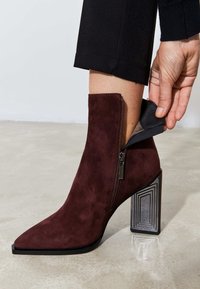 Stivale alla caviglia in suede bordeaux con punta affusolata e tacco geometrico a blocco. Dettaglio con zip sul lato per un facile accesso. Texture liscia.