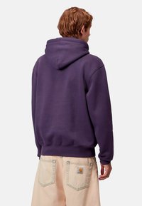 Felpa con cappuccio viola a vestibilità rilassata, con polsini e orlo a coste. I dettagli sul retro includono un logo cucito e una texture morbida.