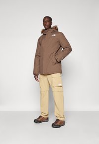 Brun isolert parka med hette trimmet med pels, beige cargo-bukser og brune tursko. Har logo detaljer og en avslappet passform.