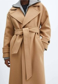 Beżowy płaszcz wełniany z paskiem w talii, szpiczastymi klapami i dwoma bocznymi kieszeniami, z szarym kapturem na bluzie pod spodem.