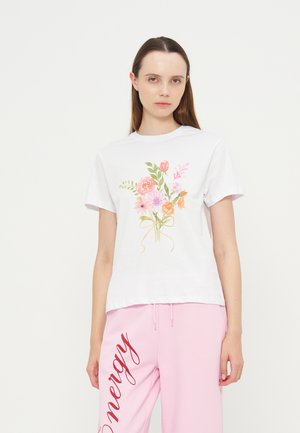 Jeune femme portant un t-shirt blanc à manches courtes avec un imprimé bouquet floral et un pantalon de survêtement rose avec un texte rouge sur une jambe.