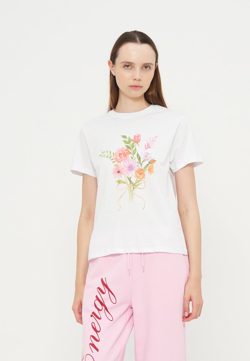 Jeune femme portant un t-shirt blanc à manches courtes avec un imprimé bouquet floral et un pantalon de survêtement rose avec un texte rouge sur une jambe.