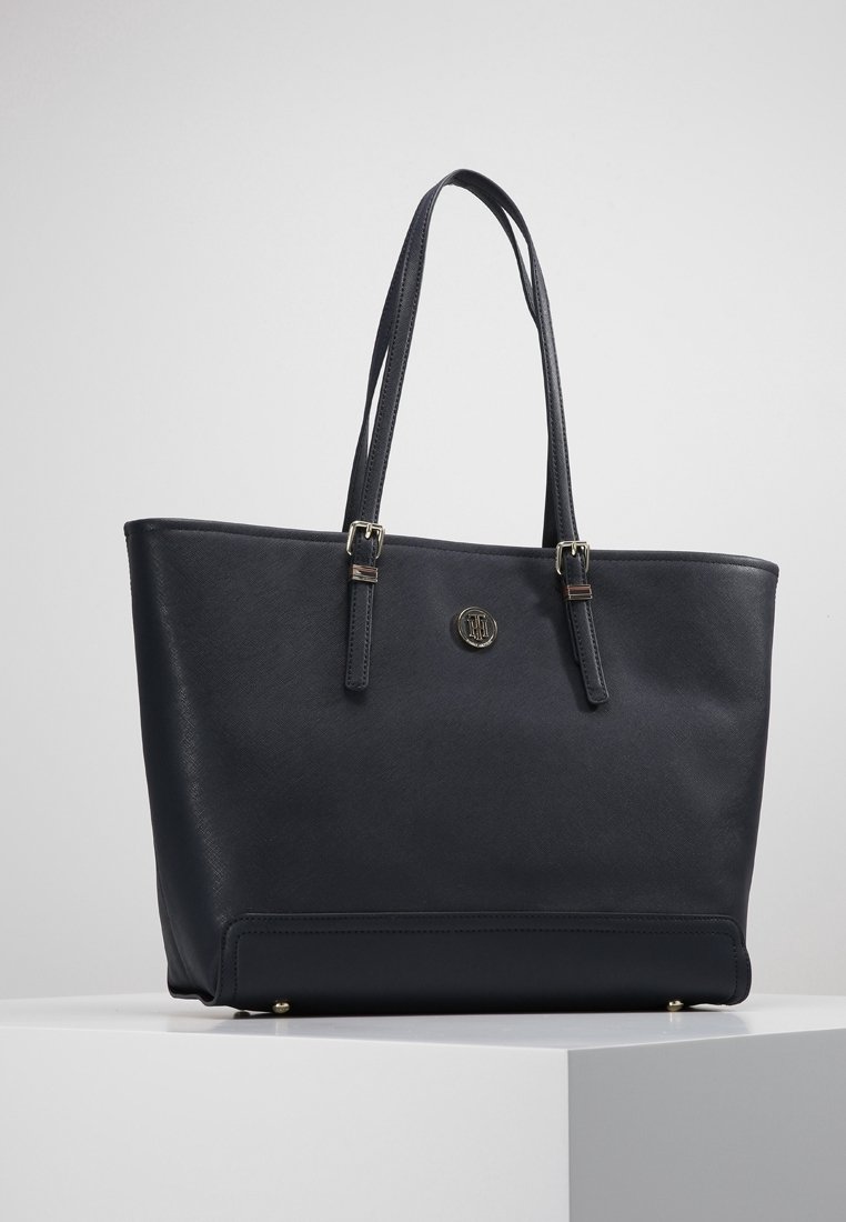 Sac cabas en cuir noir avec doubles anses, boucles dorées et un emblème rond doré à l'avant, présenté sur un socle blanc sur fond gris.