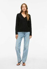 Zwarte gebreide trui met een geschulpte V-hals, gecombineerd met lichtblauwe wijde jeans en zwarte hakken. Soepele textuur, casual pasvorm.