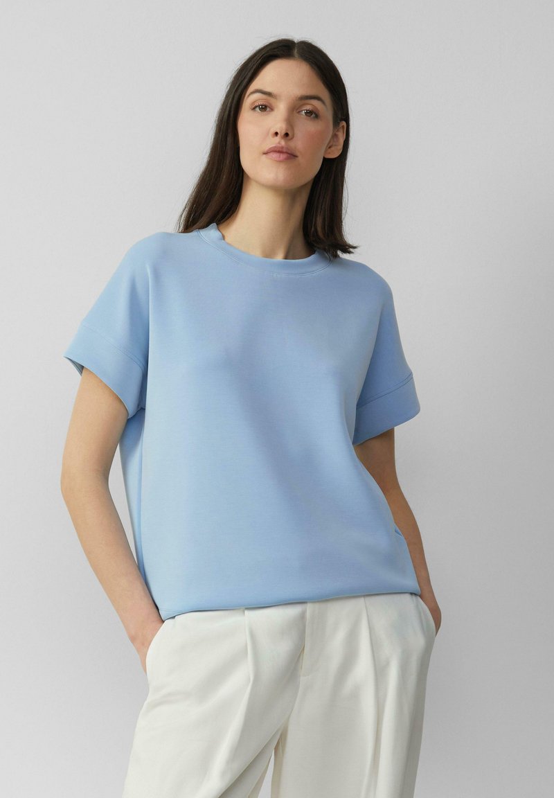 Lichtblauw T-shirt met korte, opgerolde mouwen en een ronde halslijn, gemaakt van een gladde stof. Gecombineerd met crèmewitte wijde broek.
