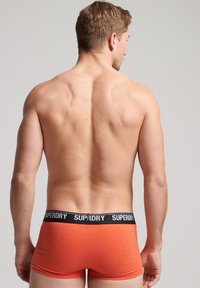 Boxer ajustados de algodón naranja con una cintura negra que presenta el logo "SUPERDRY". Vista desde atrás, resaltando el ajuste y la textura del material.