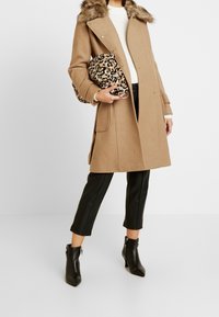 Cappotto in lana cammello con colletto foderato in pelliccia, abbinato a pantaloni aderenti neri in pelle e stivaletti neri con tacco. Tiene in mano una borsa con stampa leopardata.